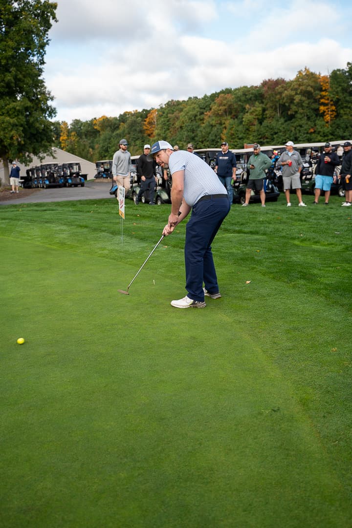 Golfer taking a shot with Classic participants gathered nearby.